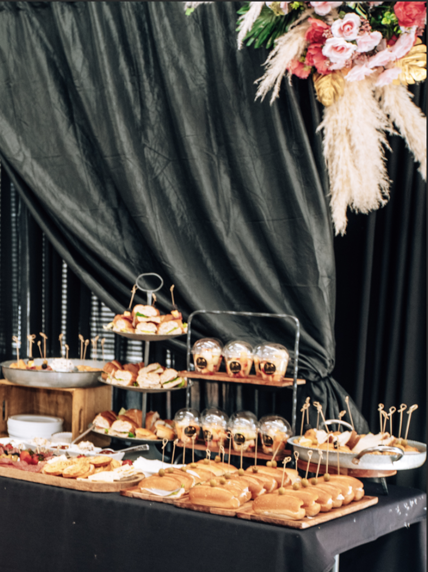 A dessert table with sandwiches, small desserts, and snacks on tiered trays and wooden boards, decorated with black drapes and a floral arrangement with pink, red, and beige flowers and pampas grass.