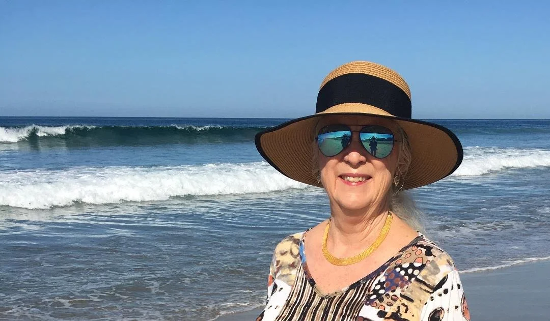 Susan Beaird wearing a large sun hat, reflective sunglasses, a patterned top, and a yellow necklace, smiling on a beach with waves and clear blue sky in the background.