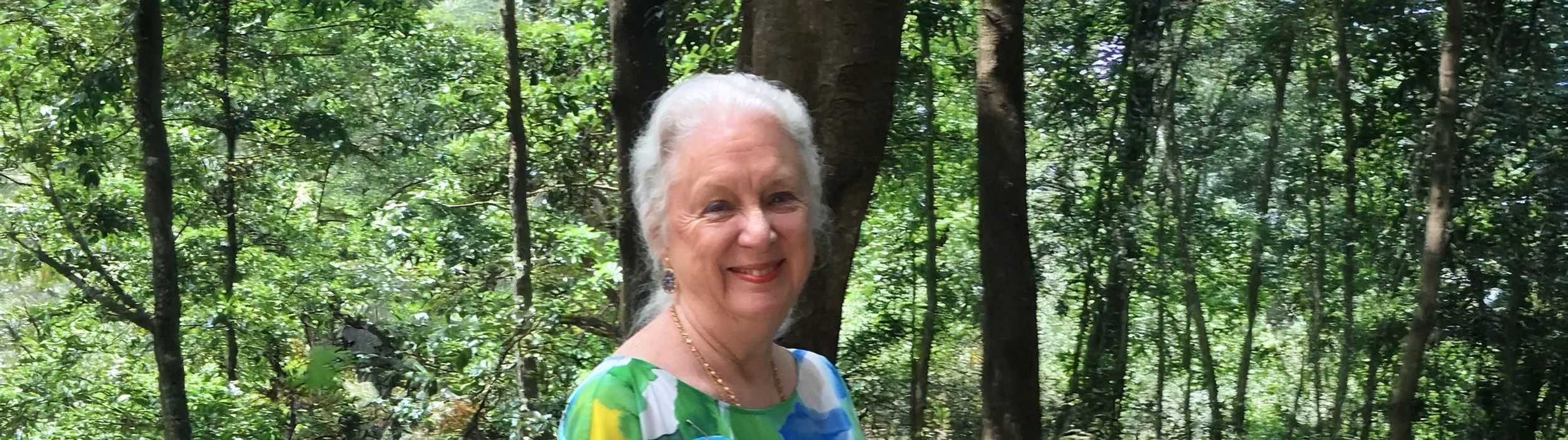 Susan Beaird with white hair smiling outdoors in a green forest, wearing a colorful blouse.