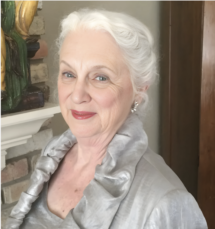 Susan Beaird with white hair styled in an updo, wearing a silver-gray satin blouse with a ruffled collar and red lipstick.