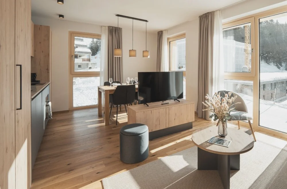 Living room with wooden flooring, large windows with curtains, a TV on a wooden stand, a dining table with chairs, a gray ottoman, a armchair, and a coffee table with a vase of flowers, in a sunny winter setting.