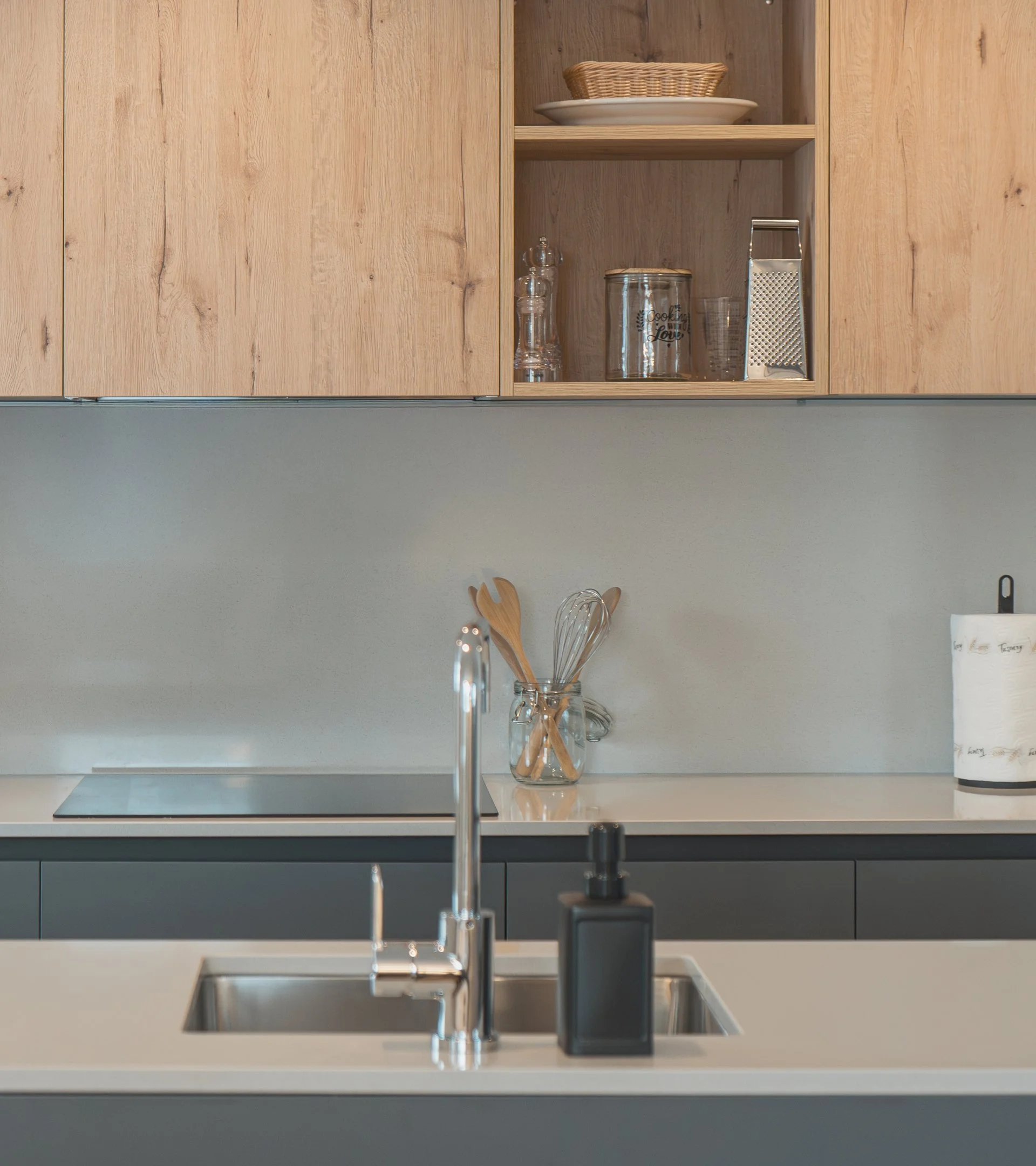 Modern kitchen with wooden upper cabinets, a sink with a faucet, and kitchen utensils in a glass jar on the counter. Open shelf with baskets and glass containers visible.