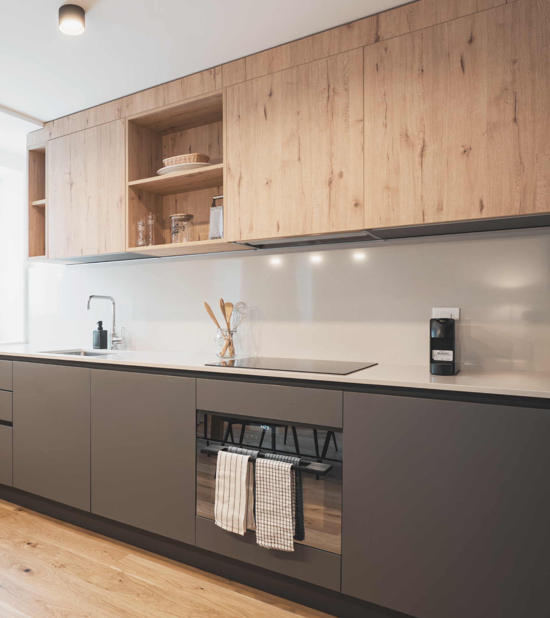 Modern kitchen with wooden upper cabinets, white backsplash, and gray lower cabinets, featuring a black induction cooktop, wine towels hanging from an oven door, and a few kitchen utensils in a glass container.