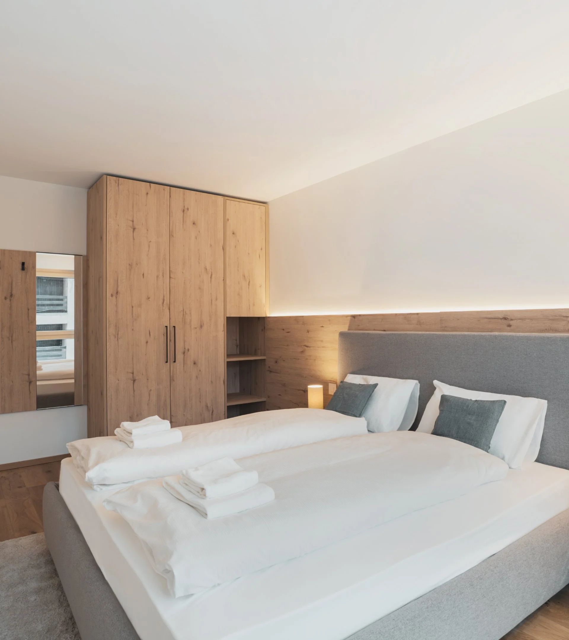 A modern hotel bedroom with a large bed, grey upholstered headboard, white linens, and three folded towels on the bed. Wooden closet and shelves are positioned behind the bed, with a small lamp and window nearby.