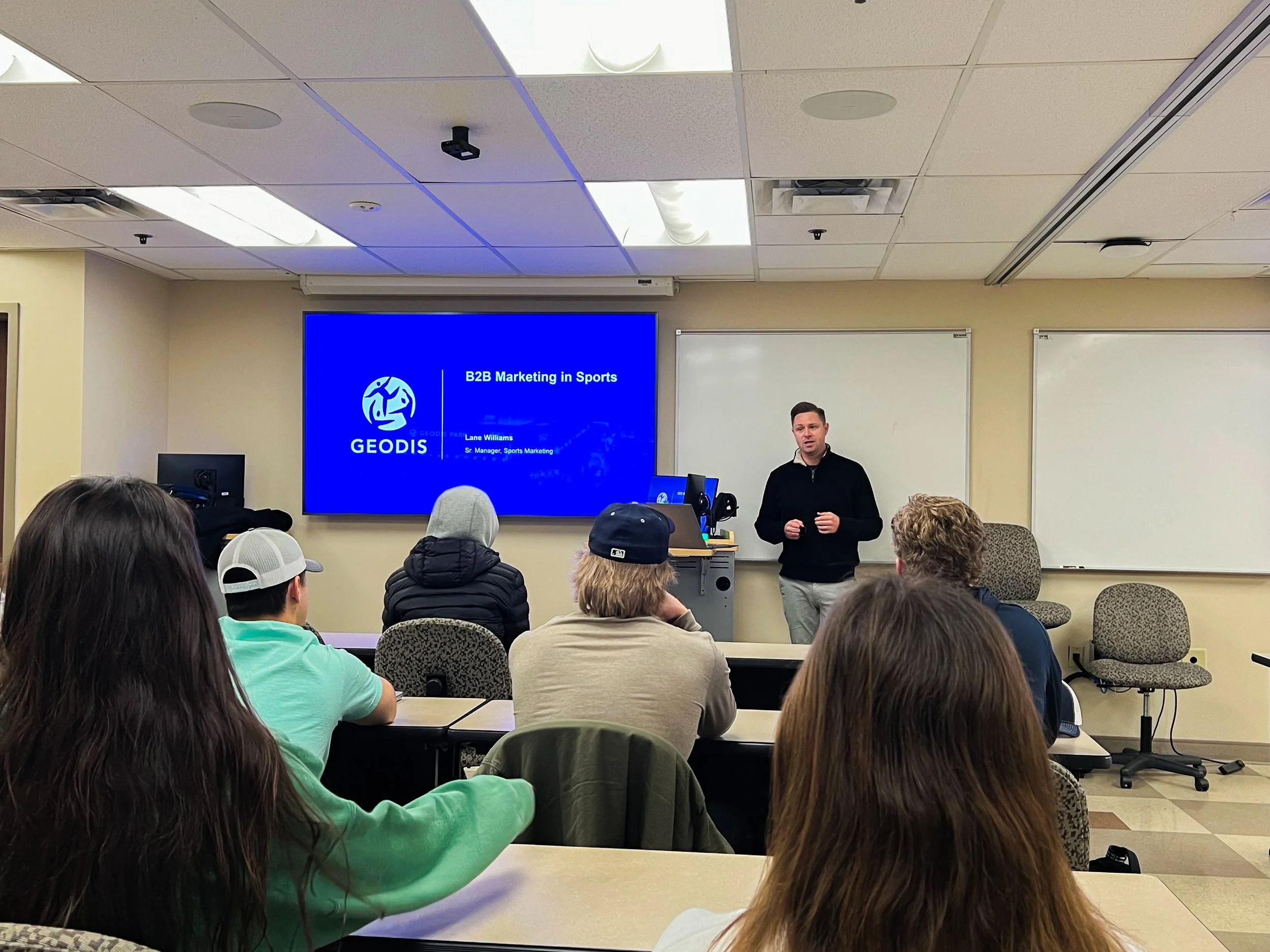 A classroom or conference room with people seated at tables, listening to a man speaking at the front. A large screen displays a presentation titled 'B2B Marketing in Sports' by GEODIS, with a logo and a subtitle mentioning Lane Williams, Senior Manager of Sports Marketing.