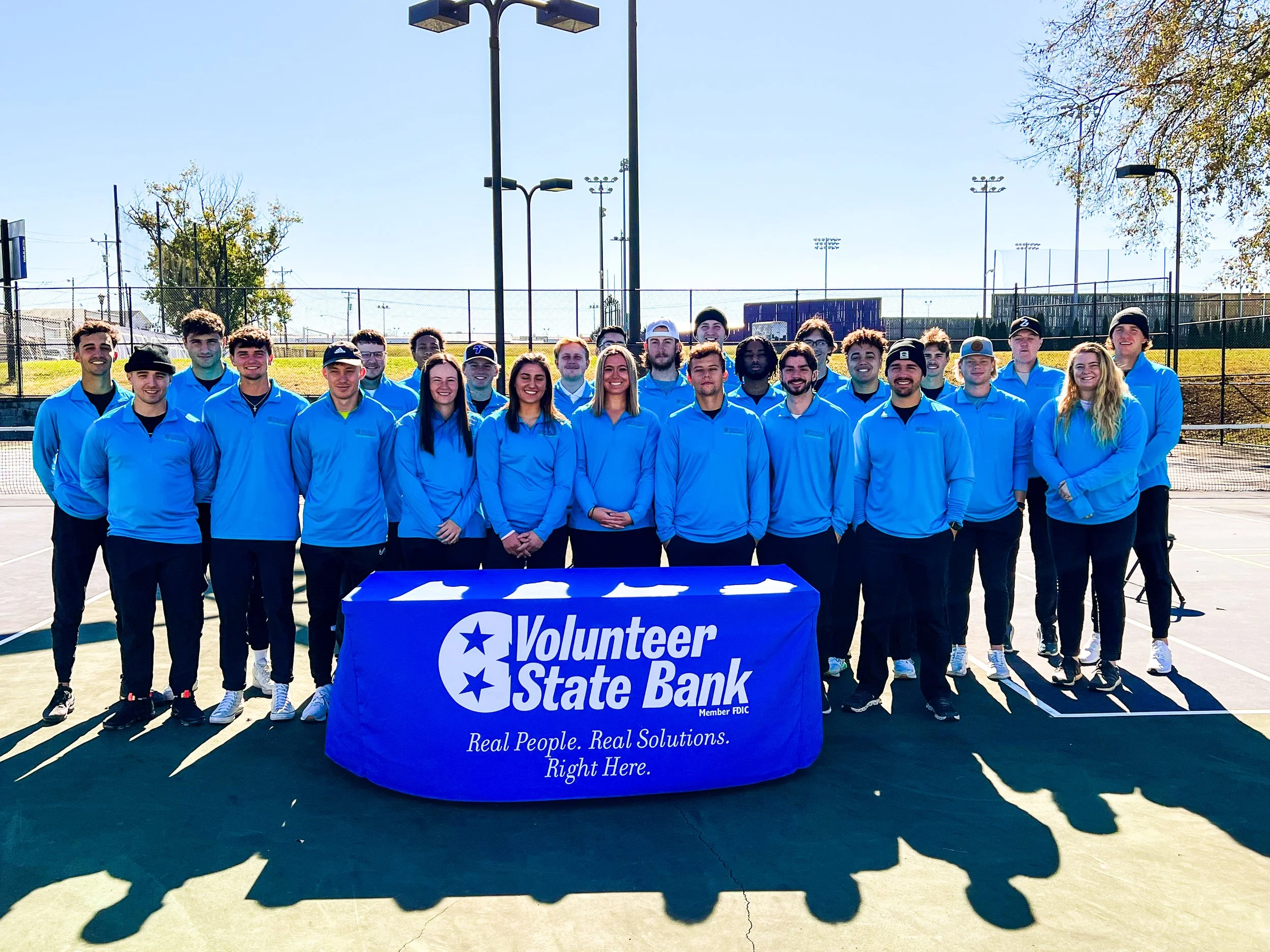 Group of Trevecca Students in matching light blue jackets standing on a tennis court behind a table with a 'Volunteer State Bank' banner, for a pickleball tournament.