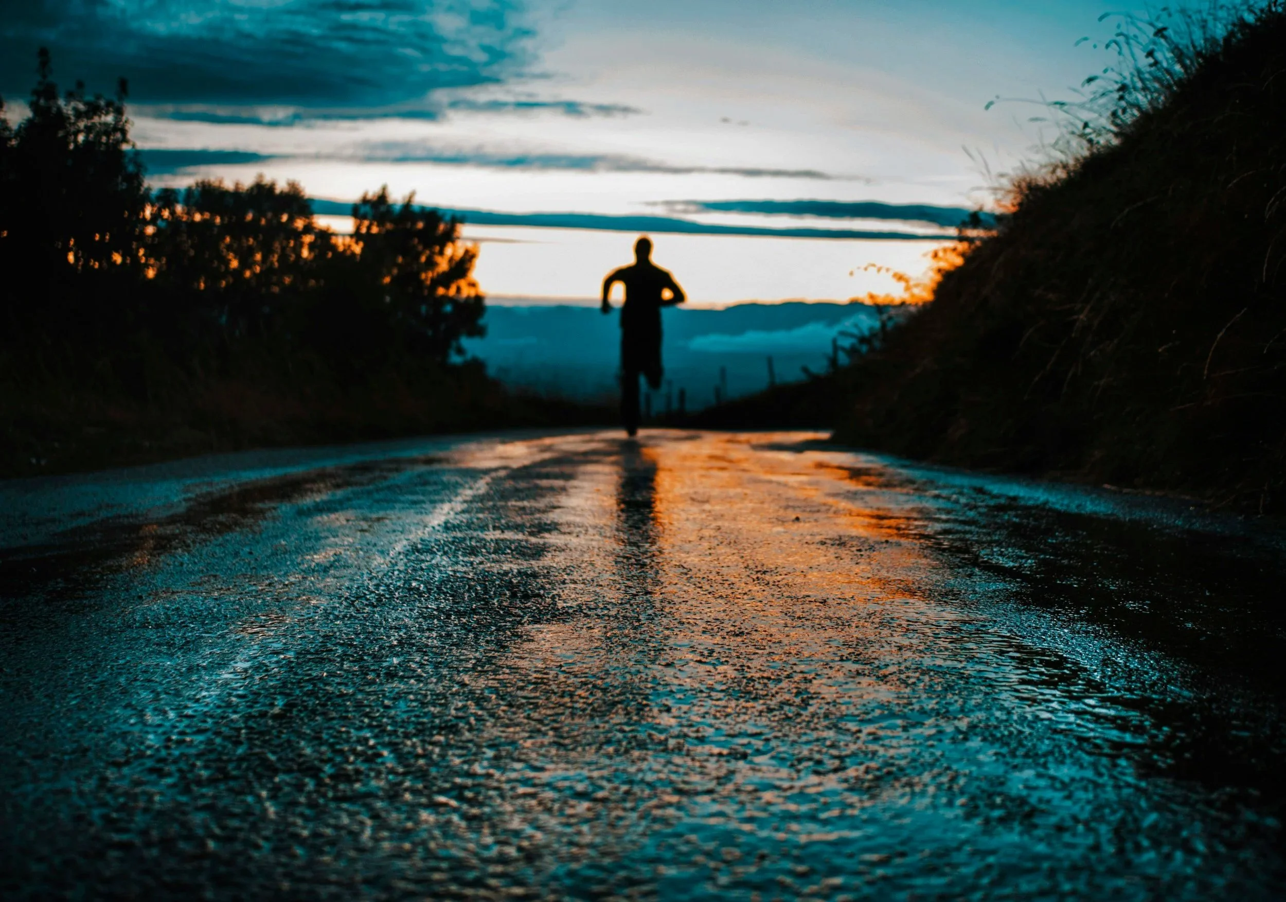 A person jogging on a wet, reflective road during sunset, with trees and hills on either side.