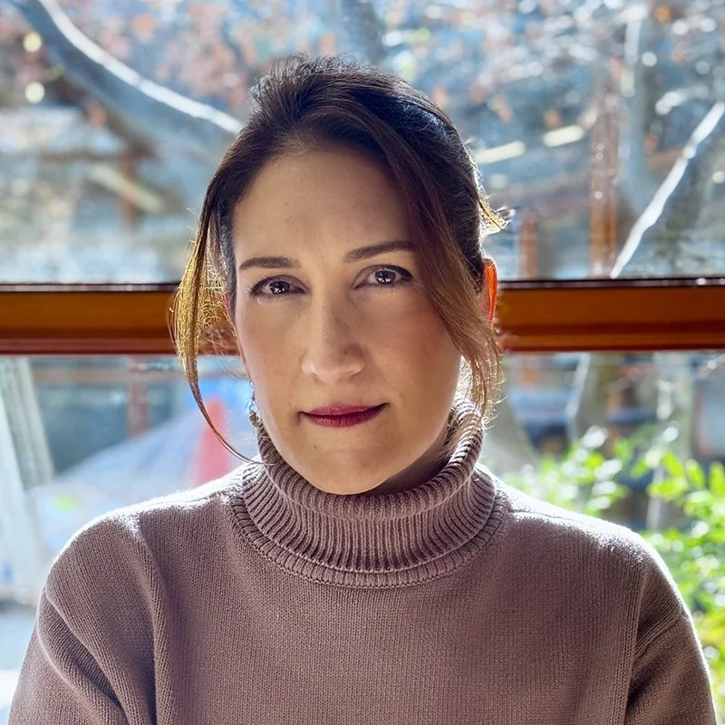 A woman sitting indoors near a window with a tree and sky visible outside. She is wearing a beige turtleneck sweater and has dark hair pulled back.