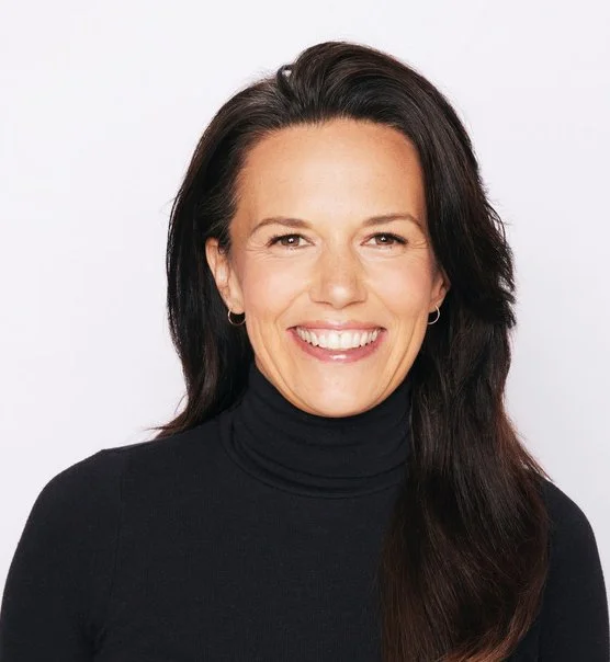 A woman with dark brown hair wearing a black turtleneck, smiling against a white background.
