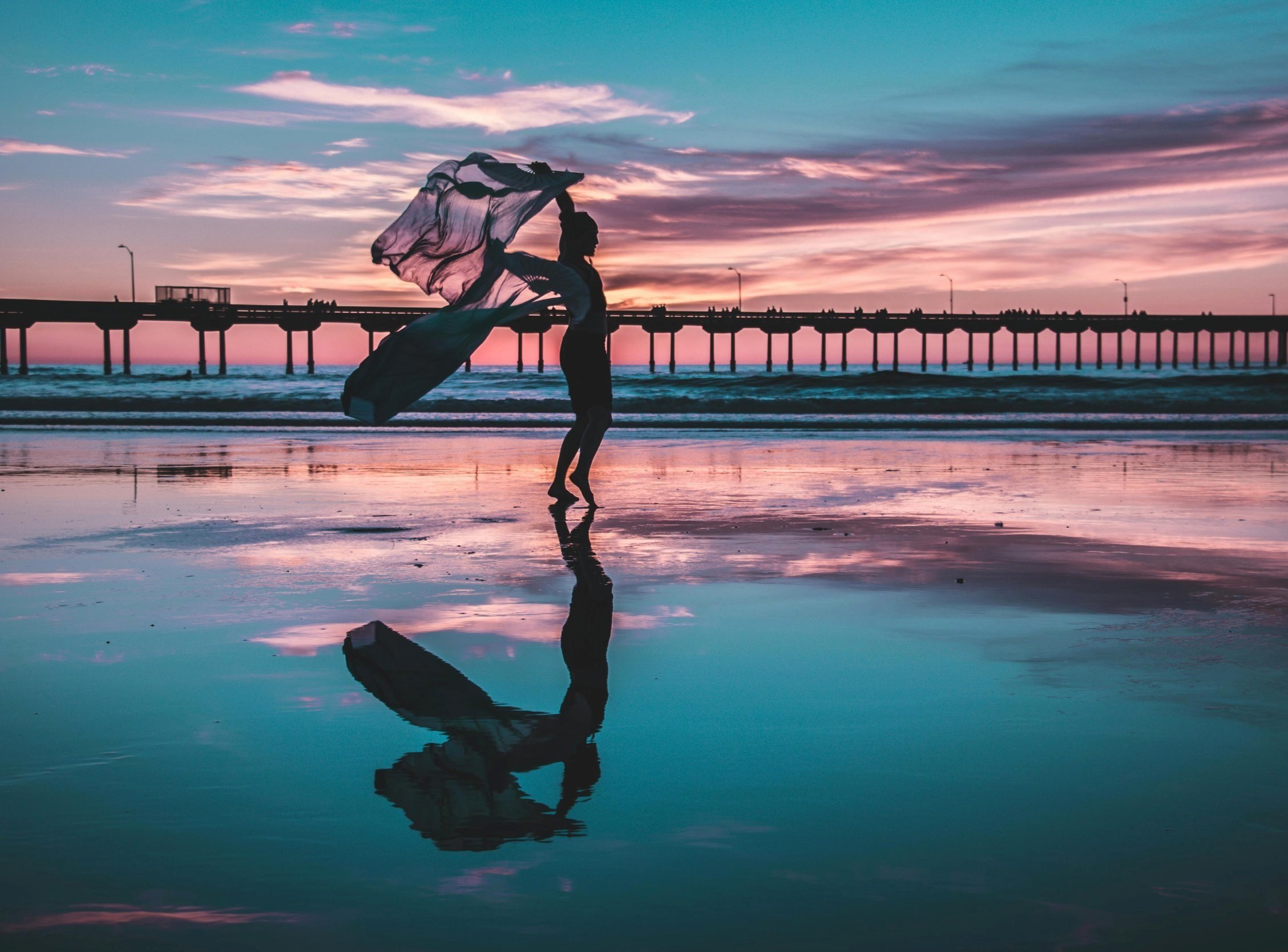 Person holding fabric on beach at sunset with pier in background, reflection in water.