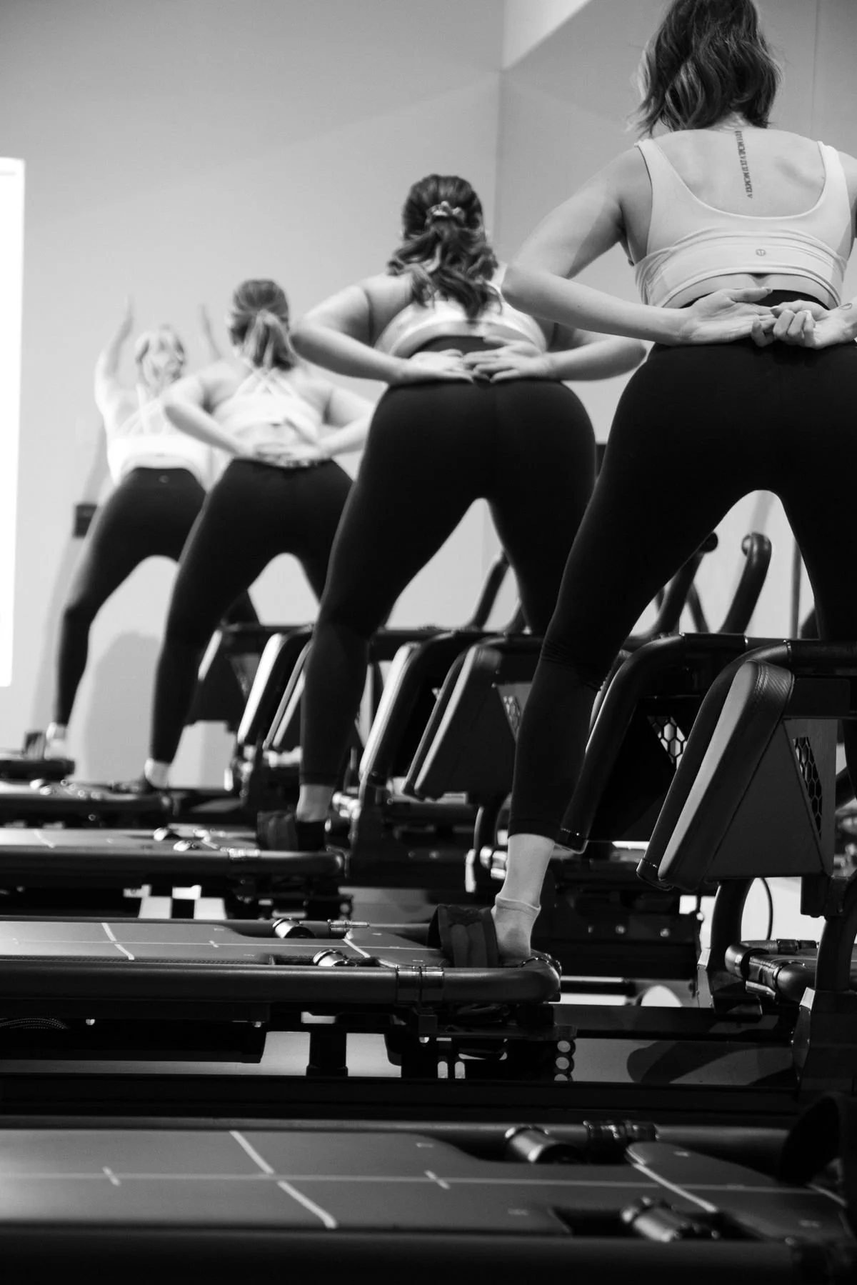 Three women in workout clothes on exercise machines in a fitness class, stretching their hands behind their backs.