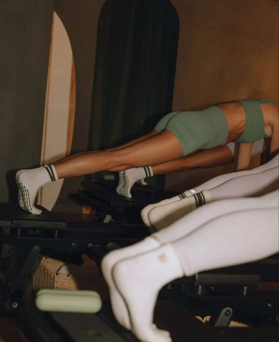 A person in athletic gear, wearing a green sports bra and shorts, is performing an exercise on a reformer machine in a fitness studio. The person is in a plank position with feet on the reformer and hands on the ground.