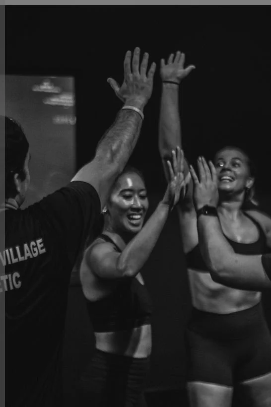 Two women with athletic build celebrating with high fives after a workout or fitness class, smiling and wearing sportswear in a gym or fitness studio.