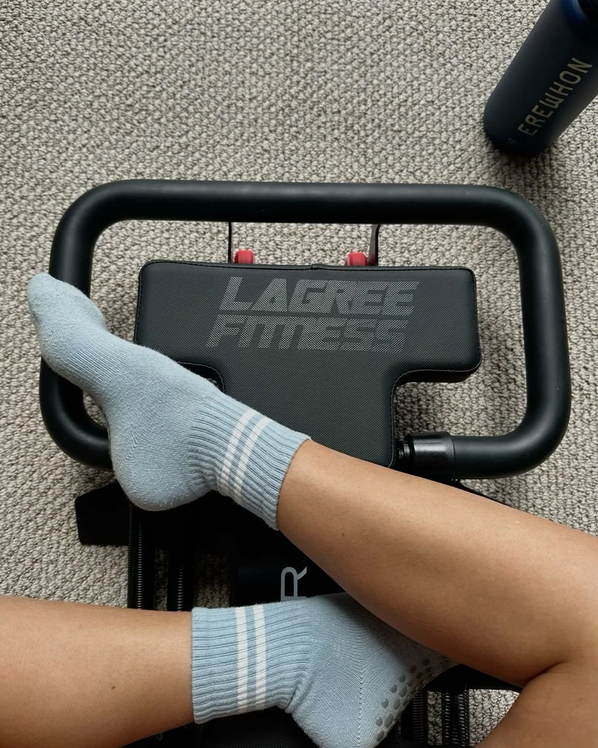Person wearing light gray socks sitting on a black exercise machine labeled 'LARGE MUSE,' on a beige carpet.