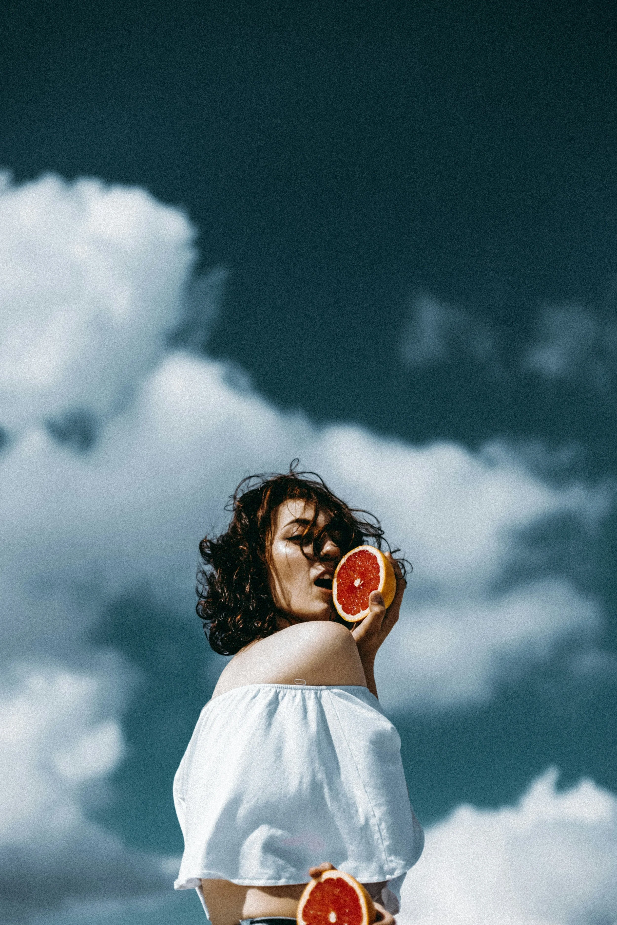 A woman with curly hair holds a half grapefruit near her face, standing outdoors with stormy clouds in the background.