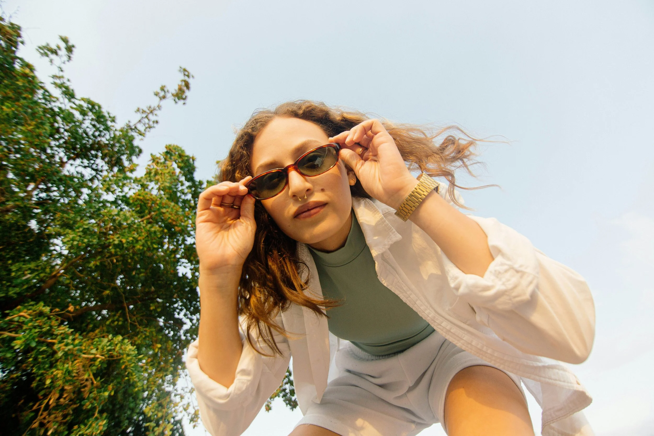 A woman with curly hair wearing sunglasses, a green top, white shorts, a beige jacket, and a gold watch, leaning forward outdoors with trees and a clear sky in the background.