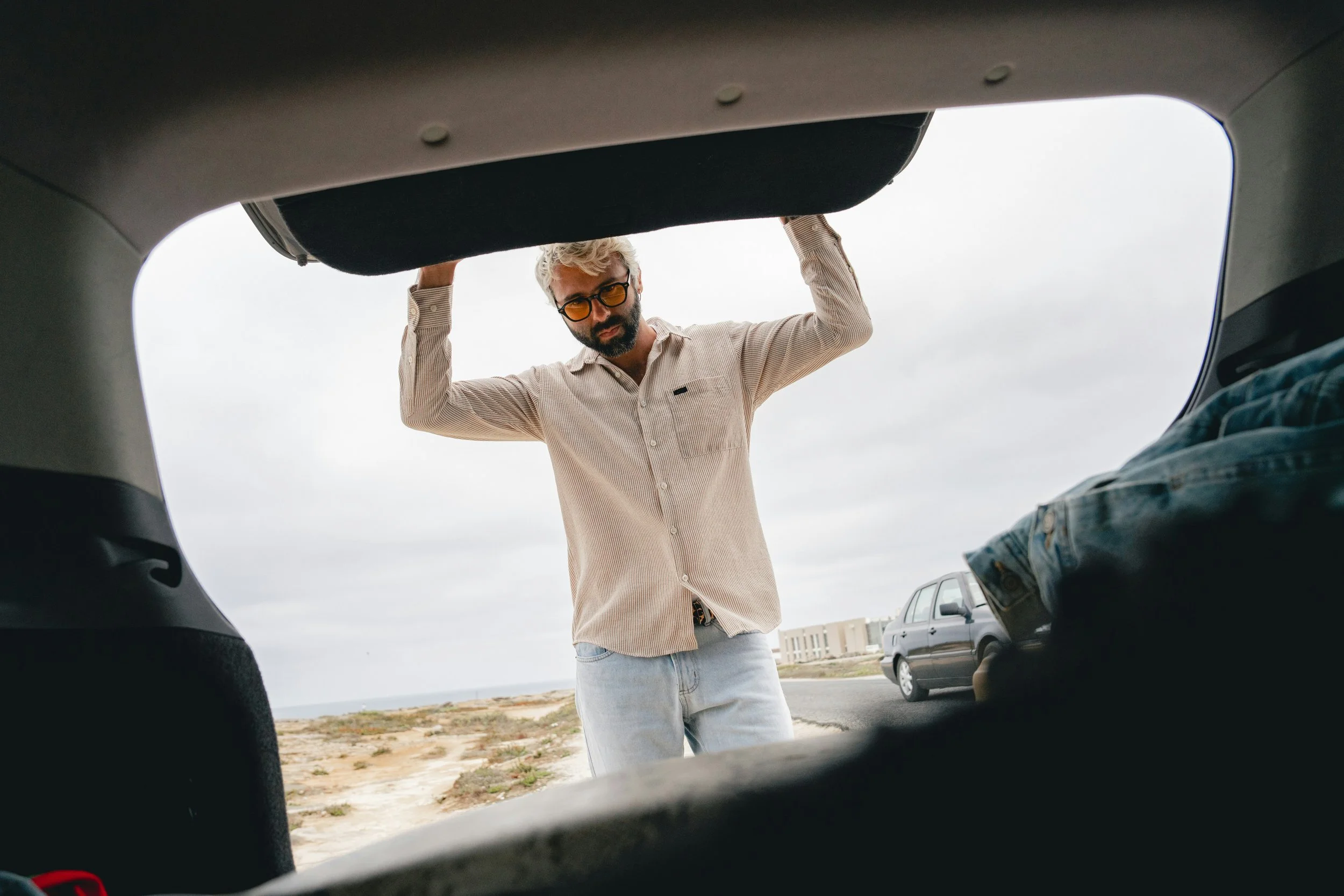 A man with sunglasses and a beard is standing outside a car, holding the trunk open with the beach and a parked SUV in the background.