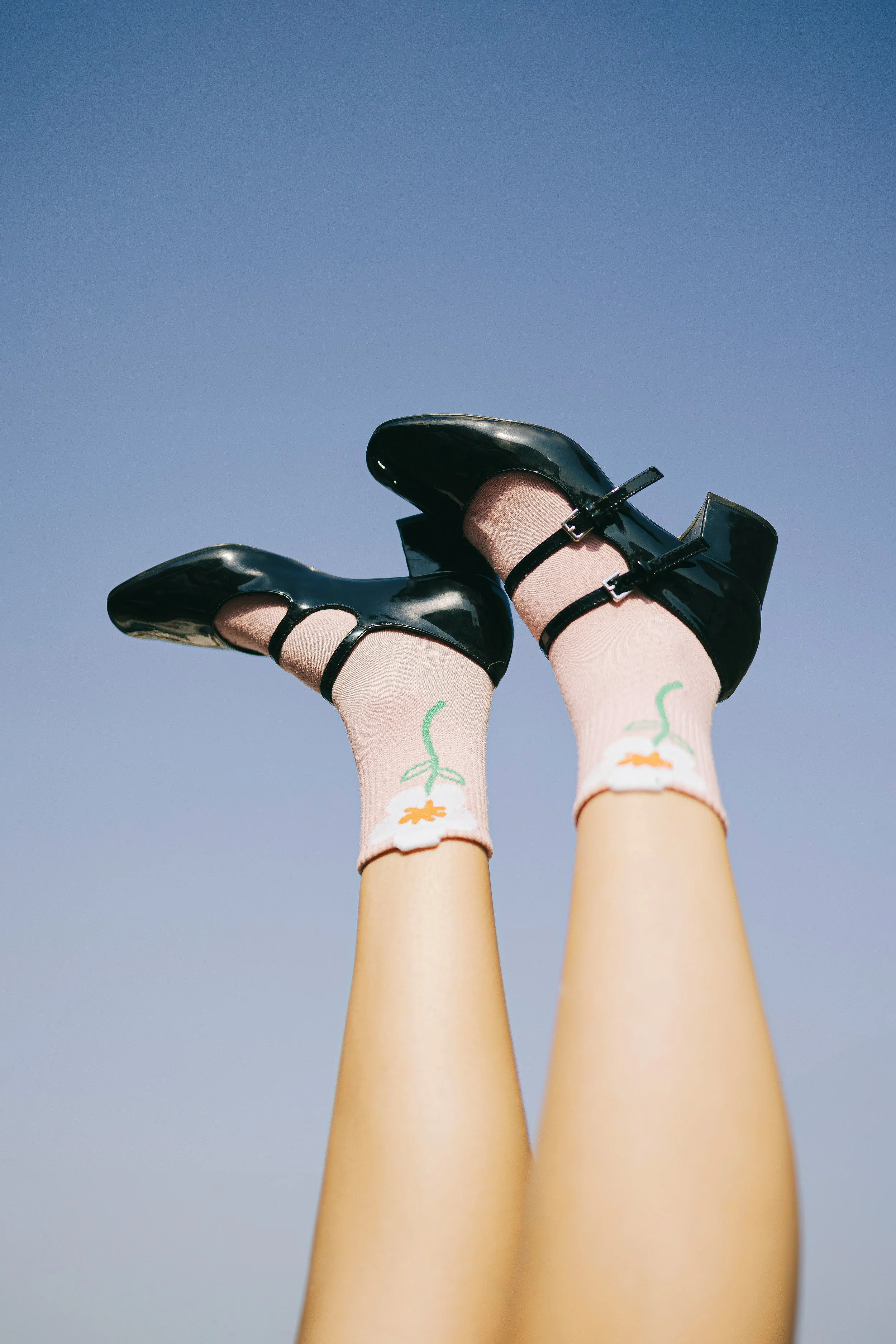A person wearing black high heels and pink socks with a white flower design, with legs raised in the air against a clear blue sky.