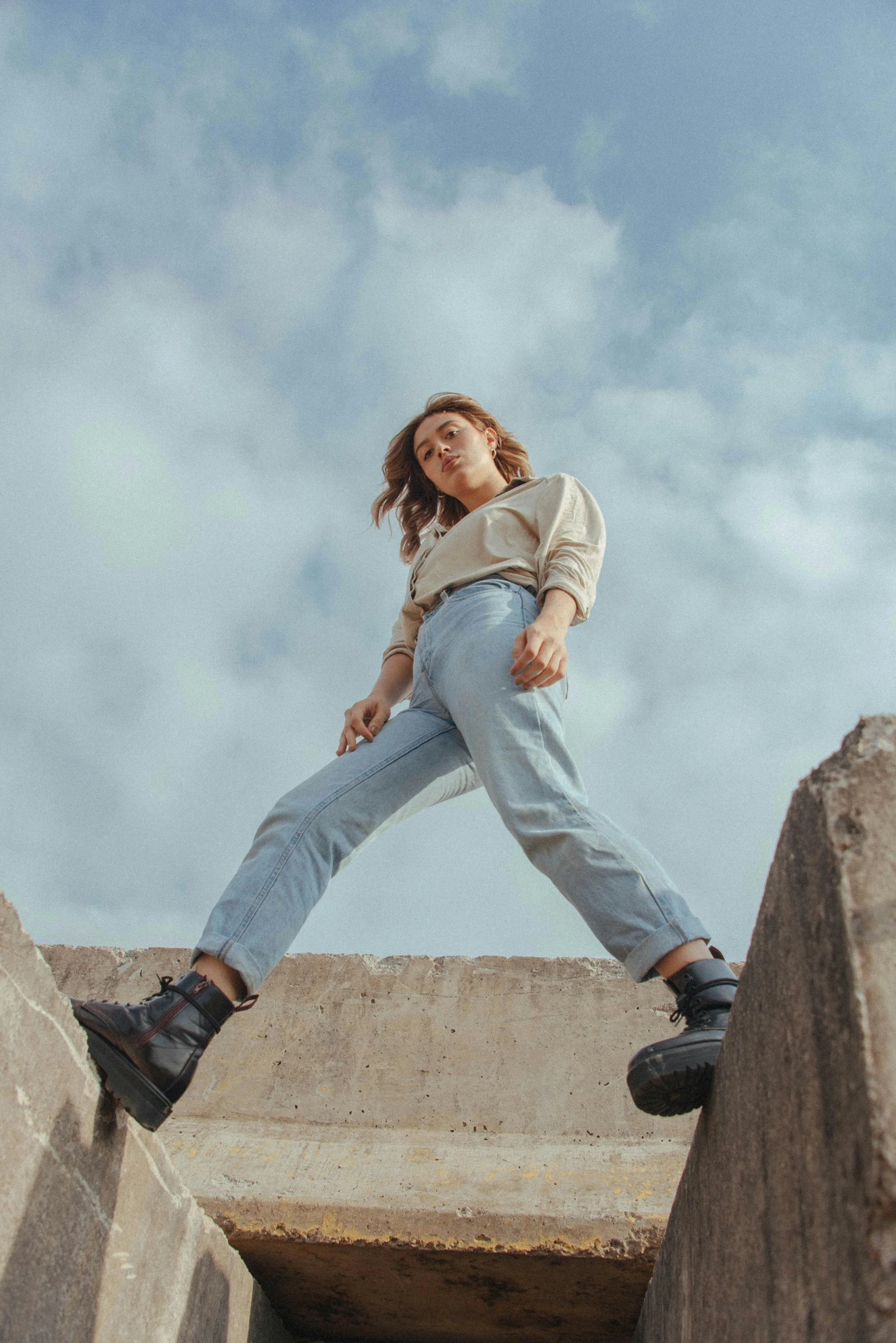 A woman standing on concrete ledges outdoors under a cloudy sky, wearing a beige shirt, light blue jeans, and black boots, looking down at the camera.