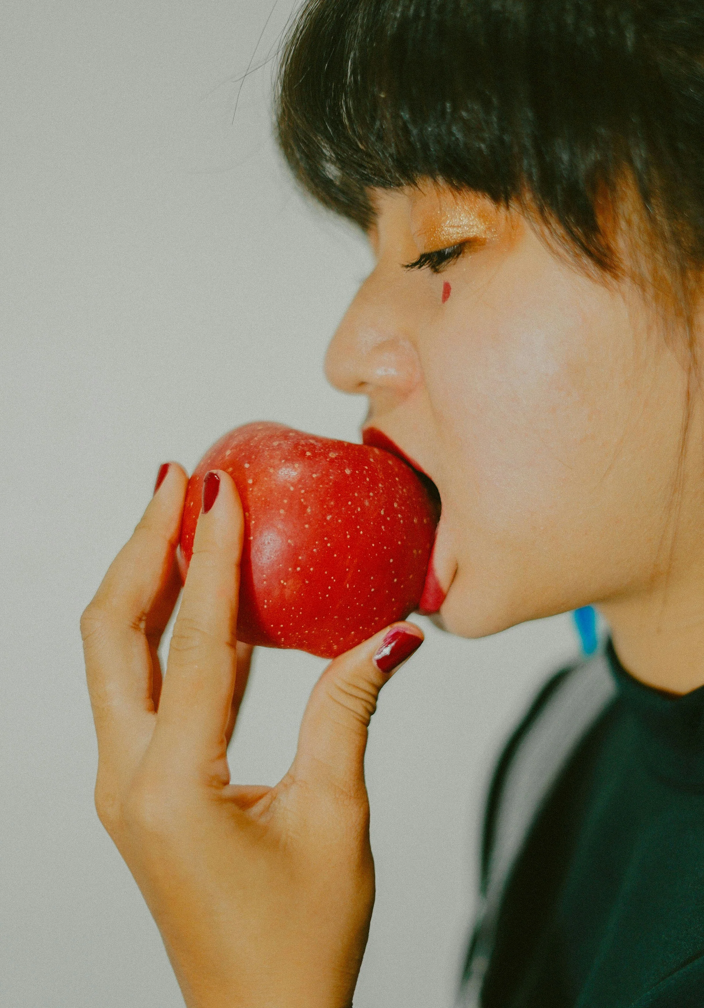 Person with dark hair and makeup biting into a red apple.