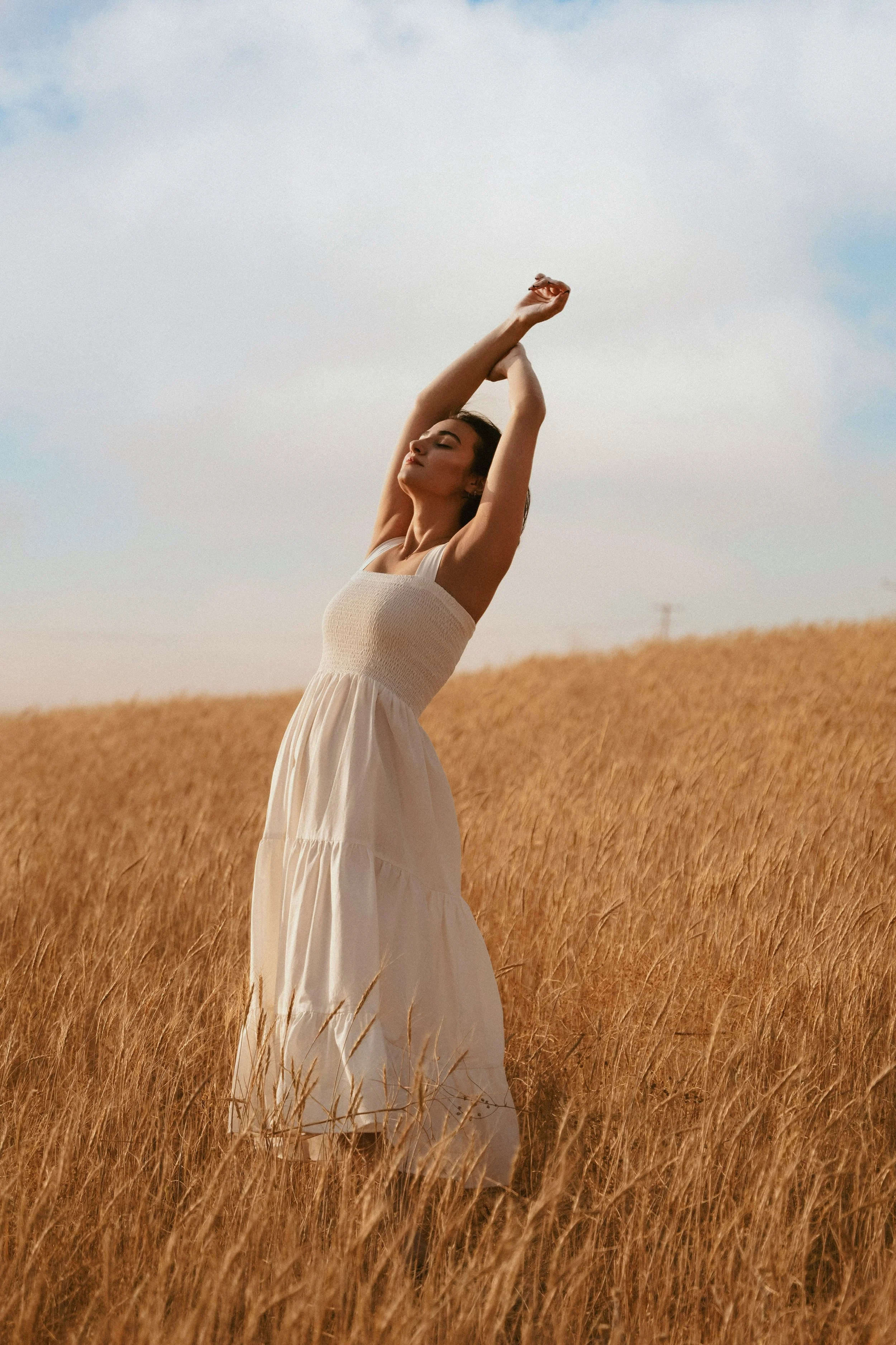 A woman in a white dress stands gracefully in a golden field of tall grass, with her arms raised above her head and eyes closed, enjoying the outdoors on a partly cloudy day.