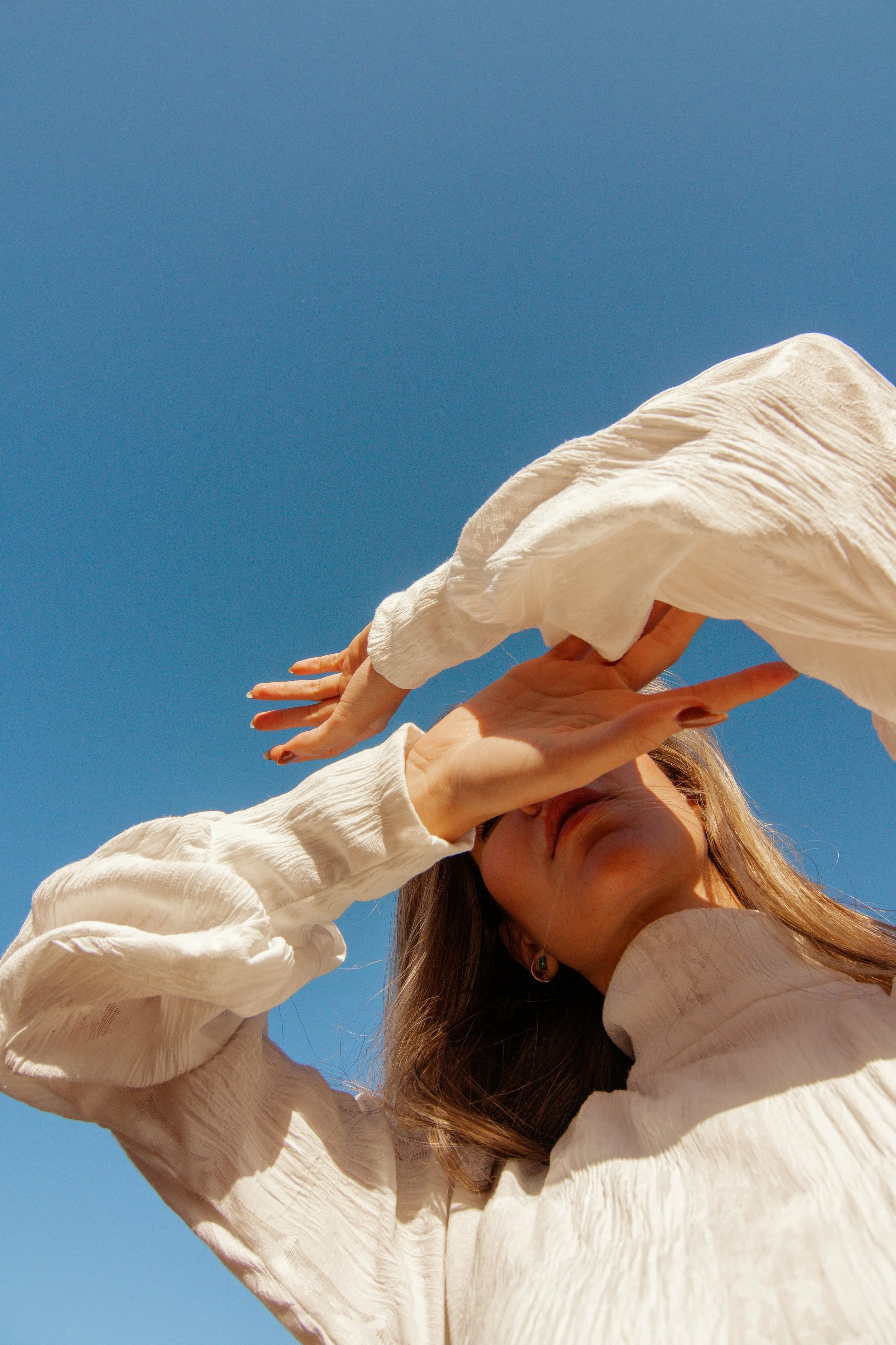 A woman in a white blouse shielding her eyes from the sun against a clear blue sky.