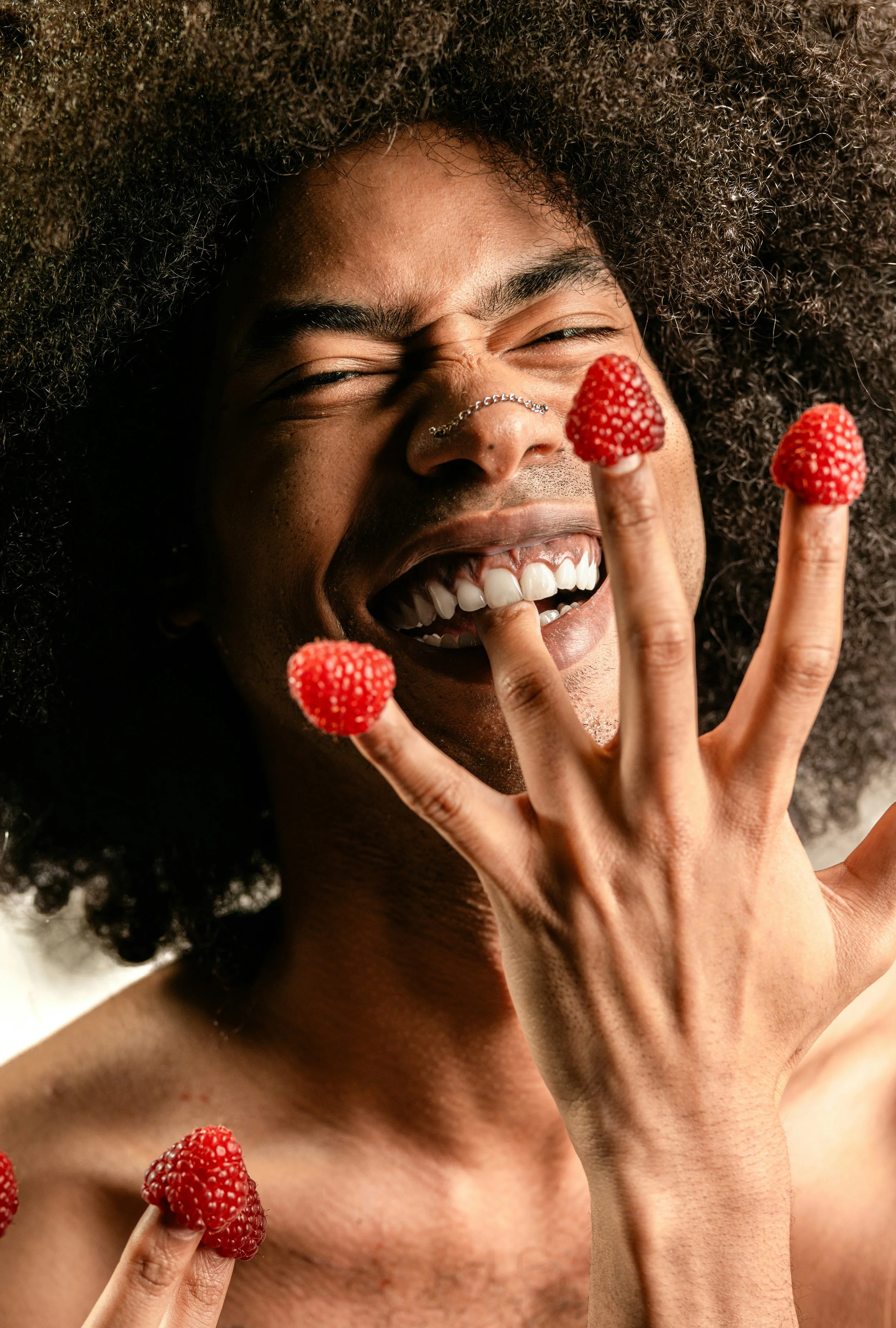 A joyful person with a large afro hairstyle, smiling with long teeth, holding a raspberry on each finger and another raspberry near their shoulder.