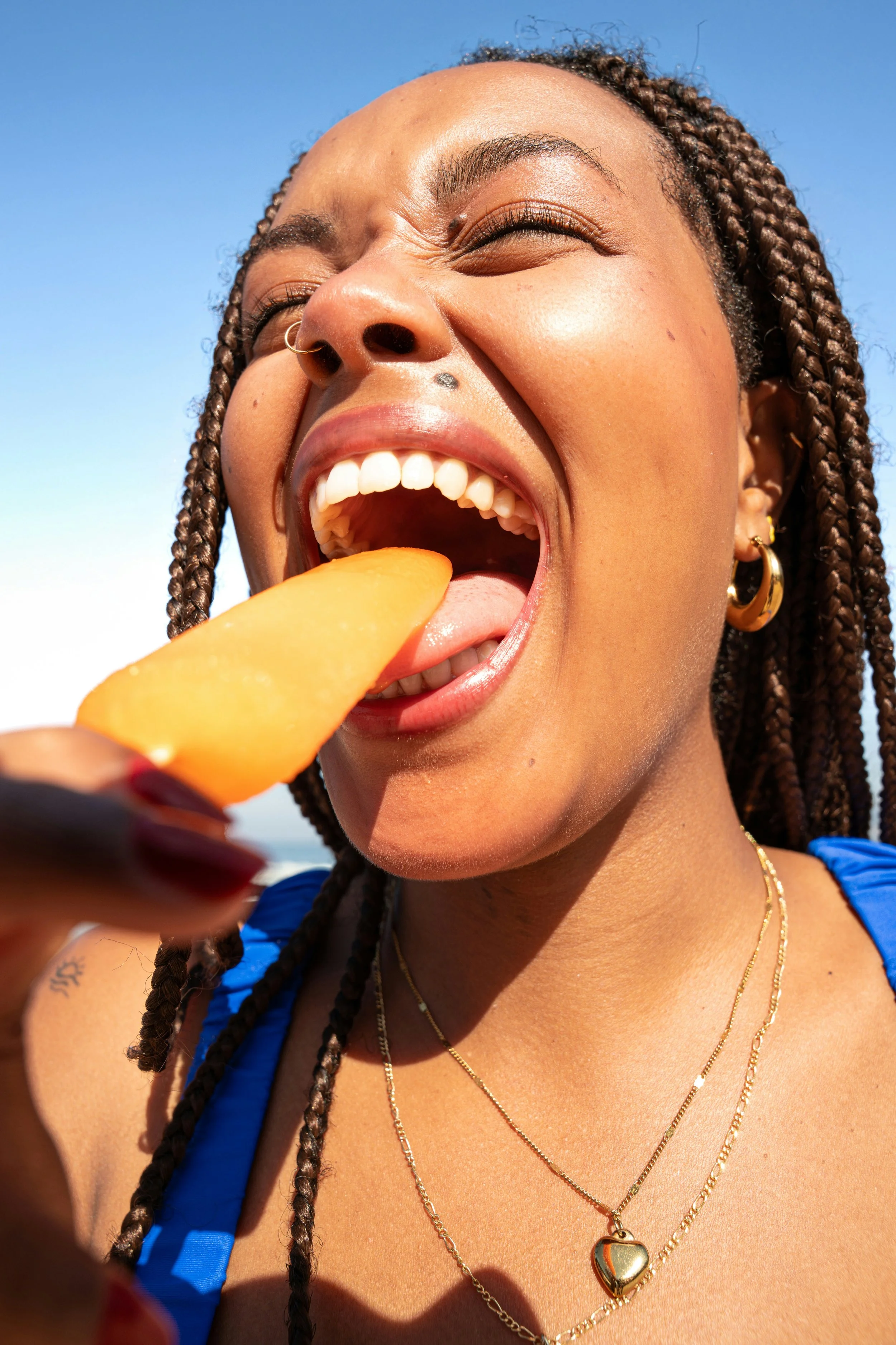 A woman with braided hair wearing a blue top, gold earrings, and layered gold necklaces, licking a mango slice with her mouth open in bright sunlight against a blue sky.