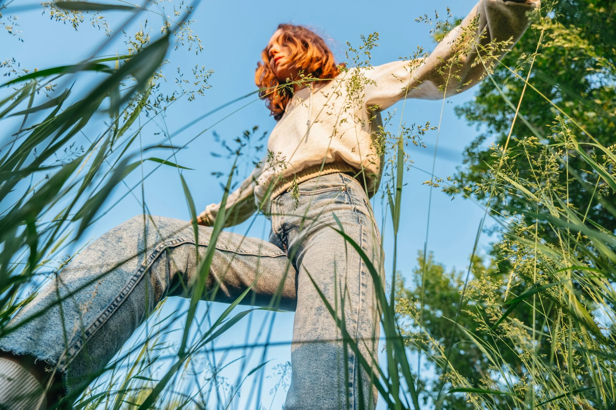 Low-angle view of a woman with red hair standing in tall grass on a sunny day, looking away from the camera.