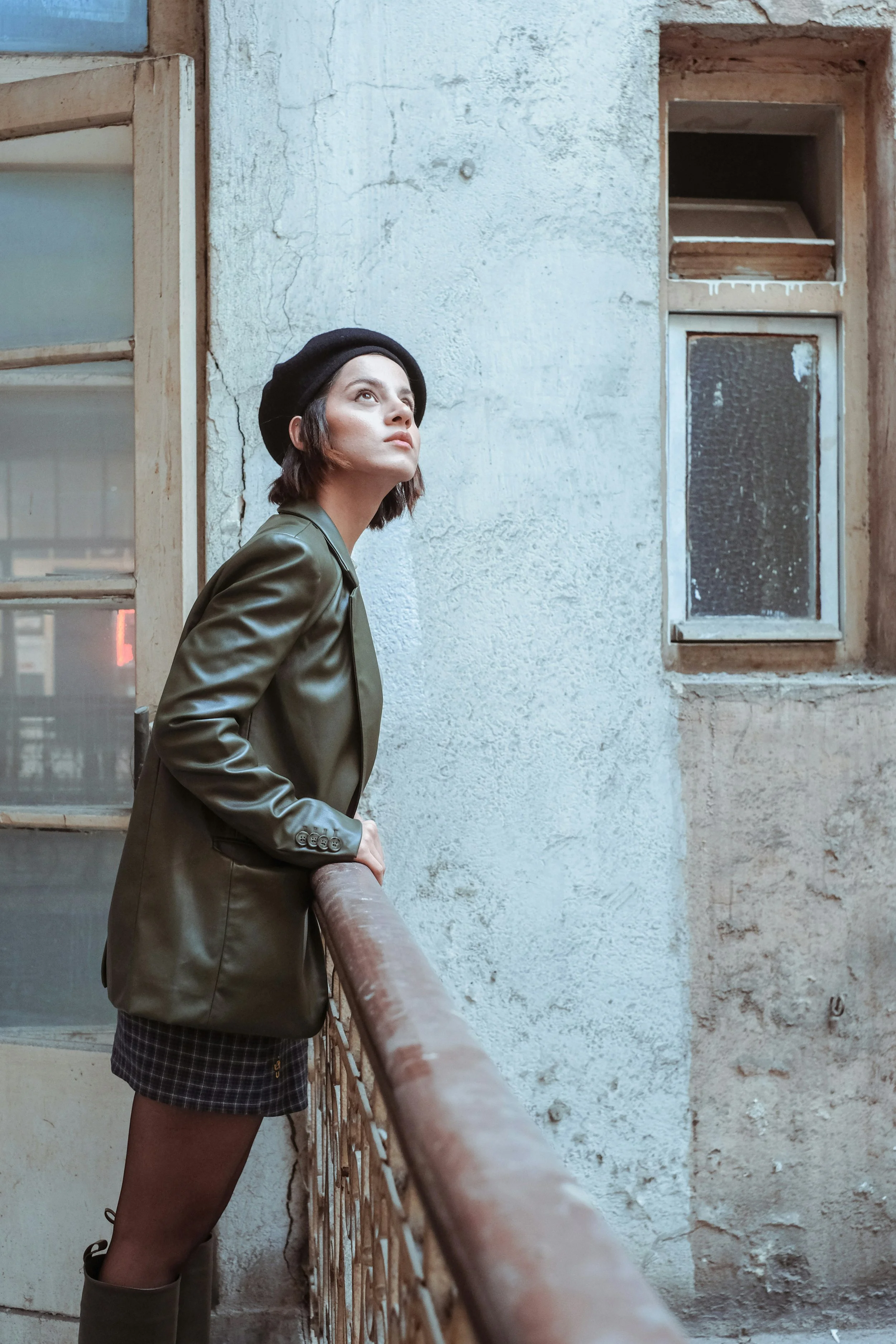 A woman with short dark hair wearing a black beret, green leather jacket, plaid skirt, and green boots looking up while leaning on a rusty railing in an old building with peeling paint and weathered windows.