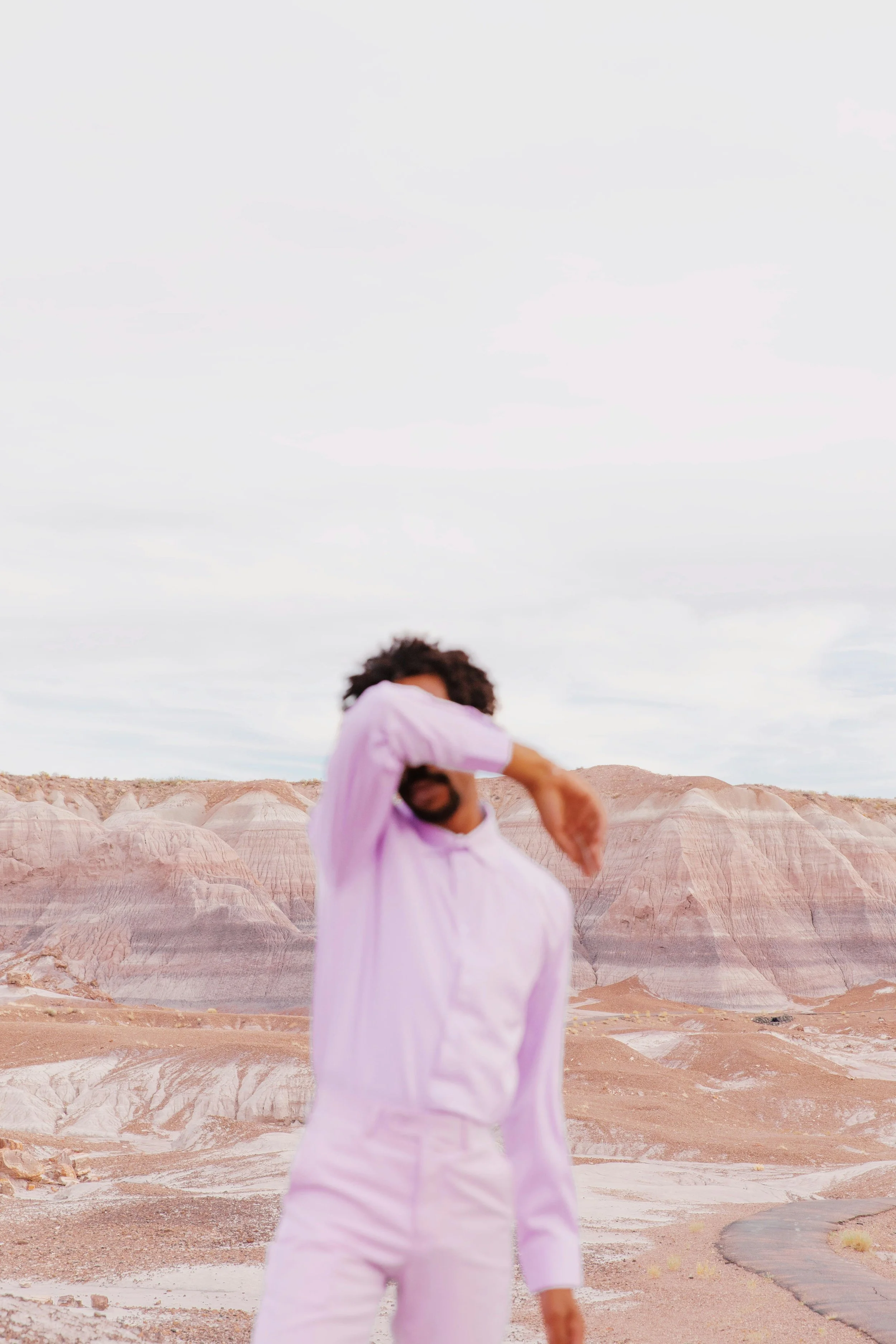 A man in a light pink shirt and pants standing in a desert landscape with multicolored hills in the background, covering part of his face with his arm.