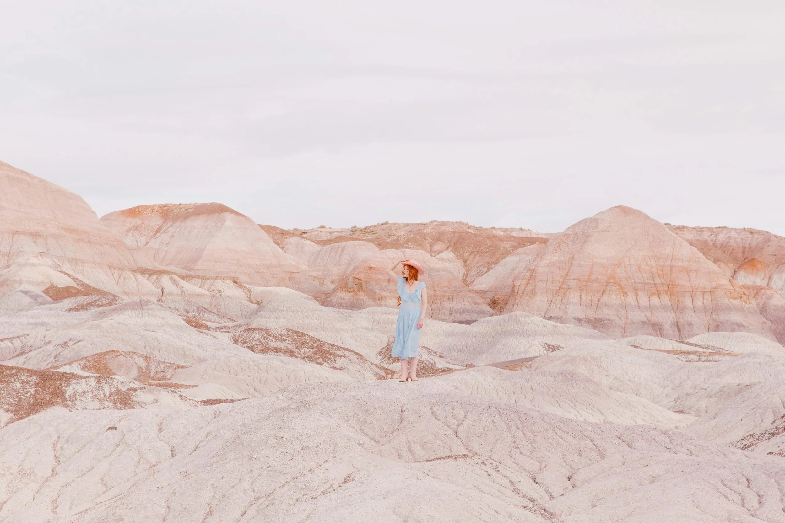 A woman in a light blue dress and wide-brimmed hat standing on a barren, rocky landscape with colorful layered hills in the background.