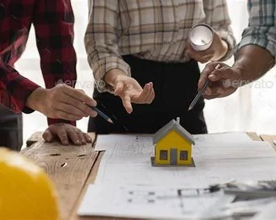 Three people examining a small model house on a table with blueprints and tools.