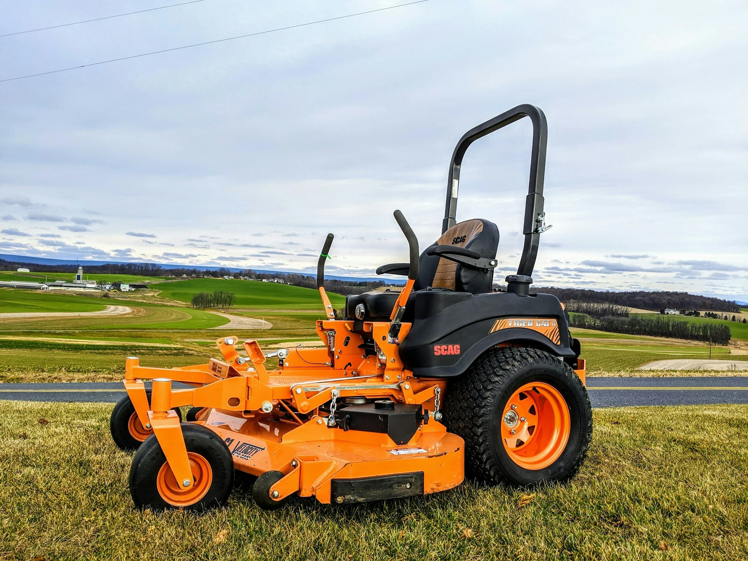 An orange and black commercial riding lawn mower on a grassy field, with rolling hills and a cloudy sky in the background.