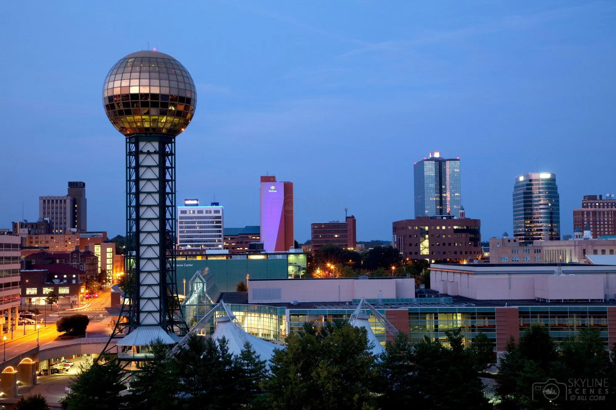 Nighttime cityscape of downtown Knoxville, Tennessee, featuring the Sunsphere with its gold-tinted glass sphere, surrounded by various lit-up modern buildings and trees in the foreground.