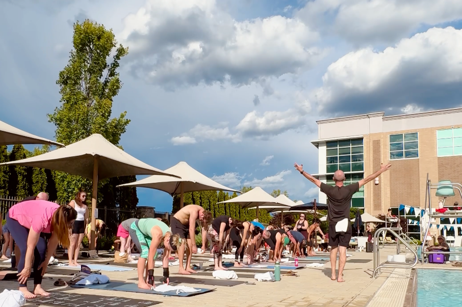 People participating in an outdoor yoga class by a swimming pool on a cloudy day, with umbrellas, trees, and a modern building in the background.