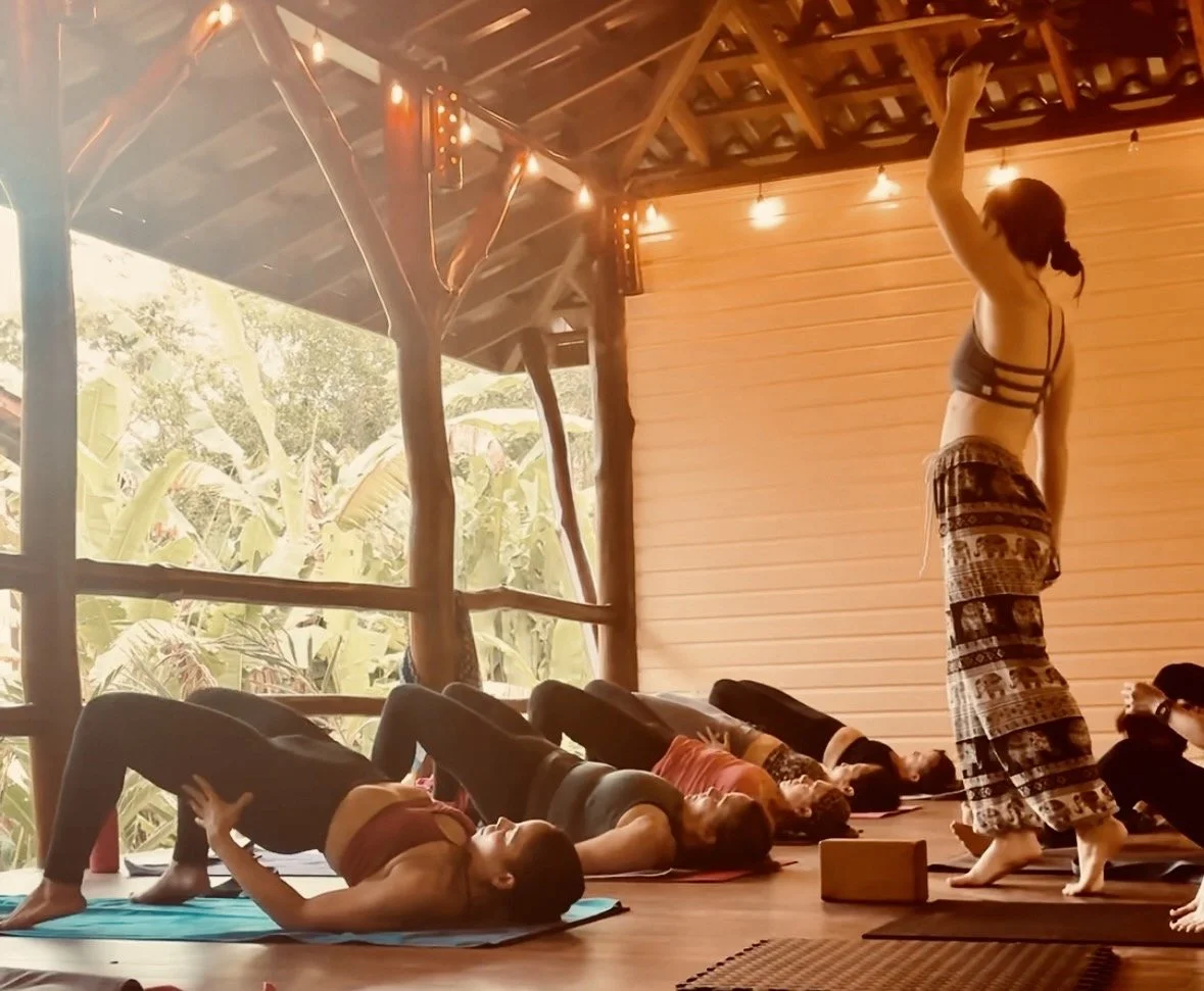 A yoga class taking place in a rustic, open-air studio in Panama with natural wood interior and string lights. The instructor is demonstrating a pose while students lie on their backs in a bridge pose on yoga mats.
