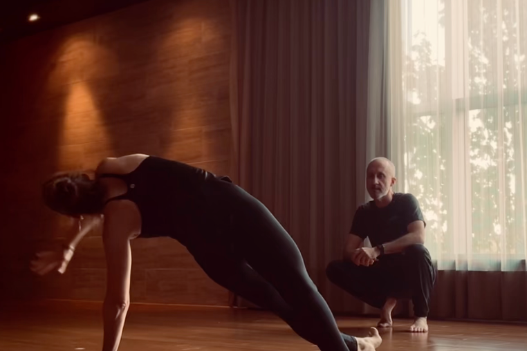 A woman in athletic clothing is doing a yoga pose on a wooden floor, with a man sitting nearby watching her, in a room with wooden walls and a large window with curtains.