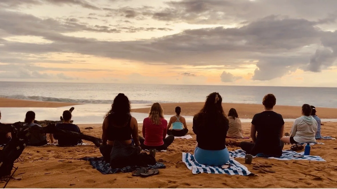People practicing yoga on the beach at sunset, sitting on towels facing the ocean with a cloudy sky overhead.