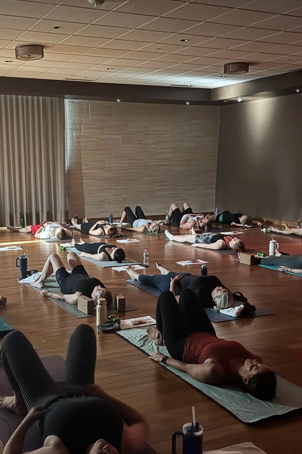 People participating in a yoga class in a spacious, dimly lit room on wooden floors, lying on mats in relaxation pose.