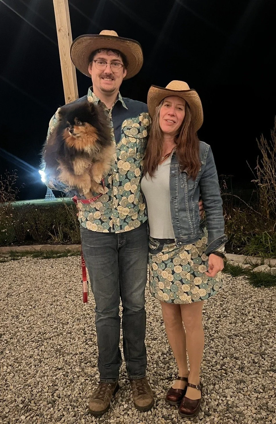 Scout and Thomas stand in complementary outfits and cowboy hats in an outdoor venue. Thomas is holding their puppy dog, JuniperNoel.