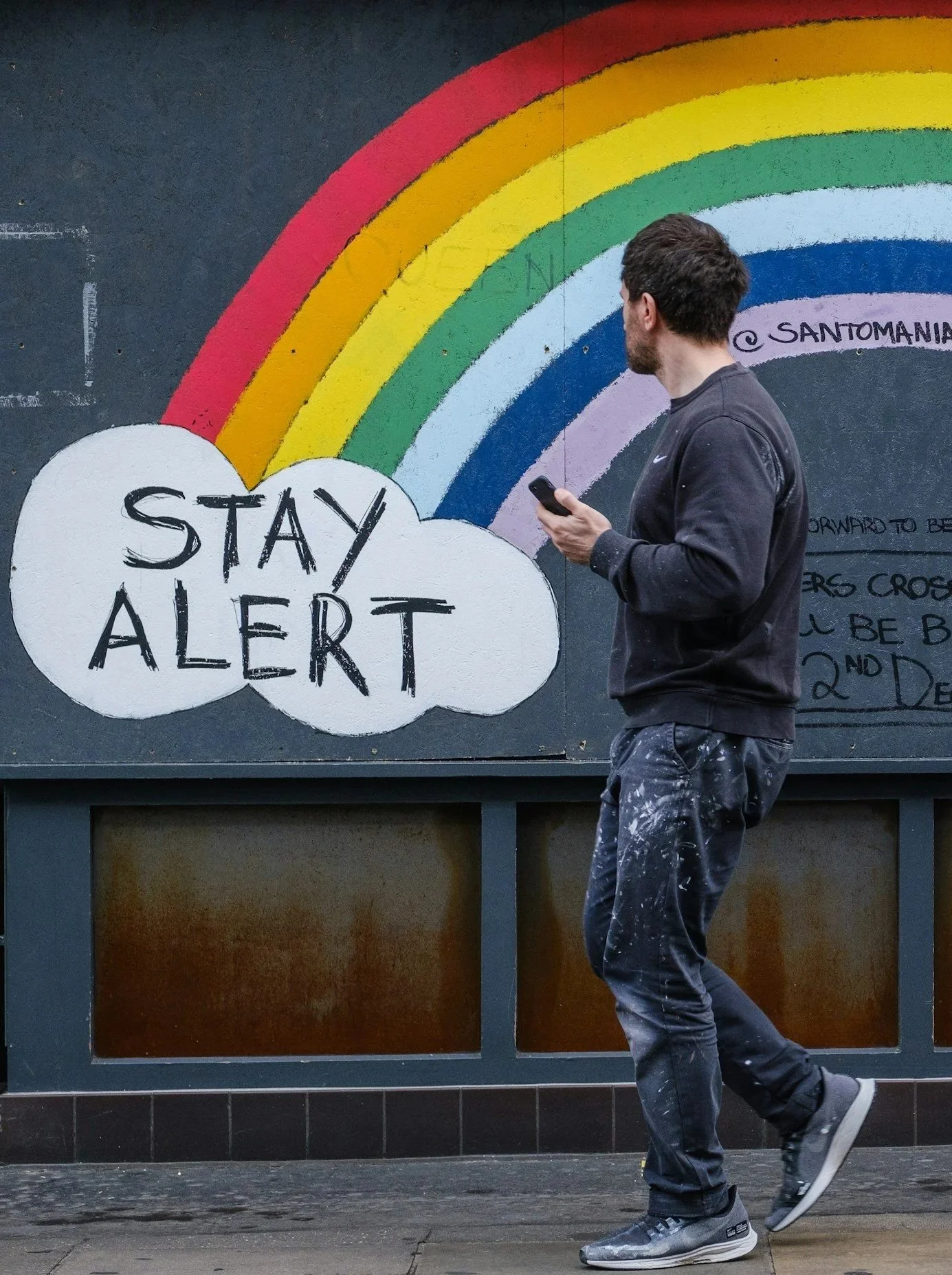 A man walks by a mural of a rainbow. In the cloud it reads "stay alert."