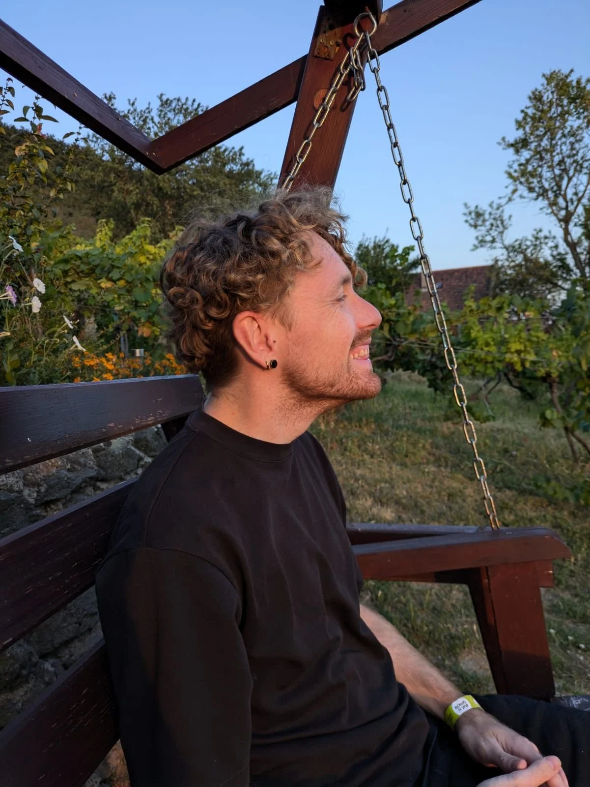 A young man with curly hair, wearing a black shirt, sitting on a wooden swing outdoors during sunset, smiling with his eyes closed.