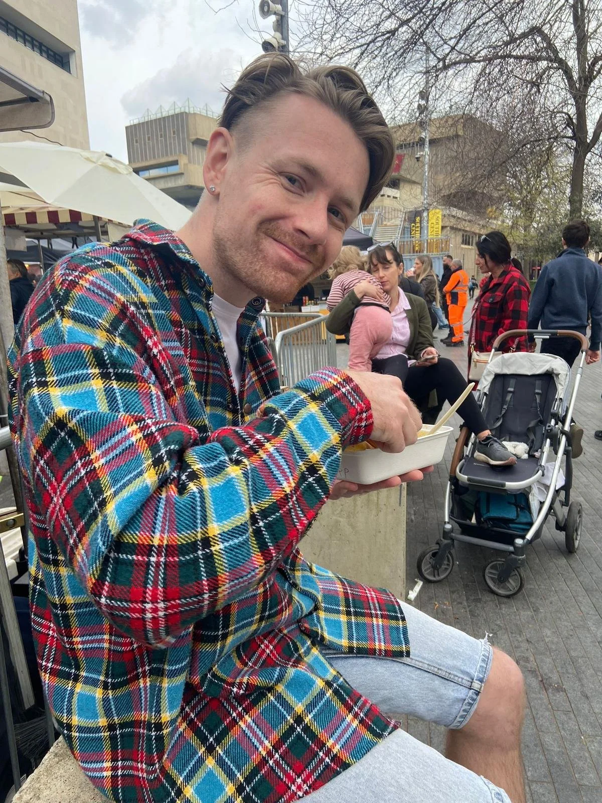 A smiling man with short hair and a beard, wearing a colorful plaid shirt, sitting outdoors at a market or fair, holding a food container and a fork. In the background, there are other people, a baby in a stroller, and market stalls.
