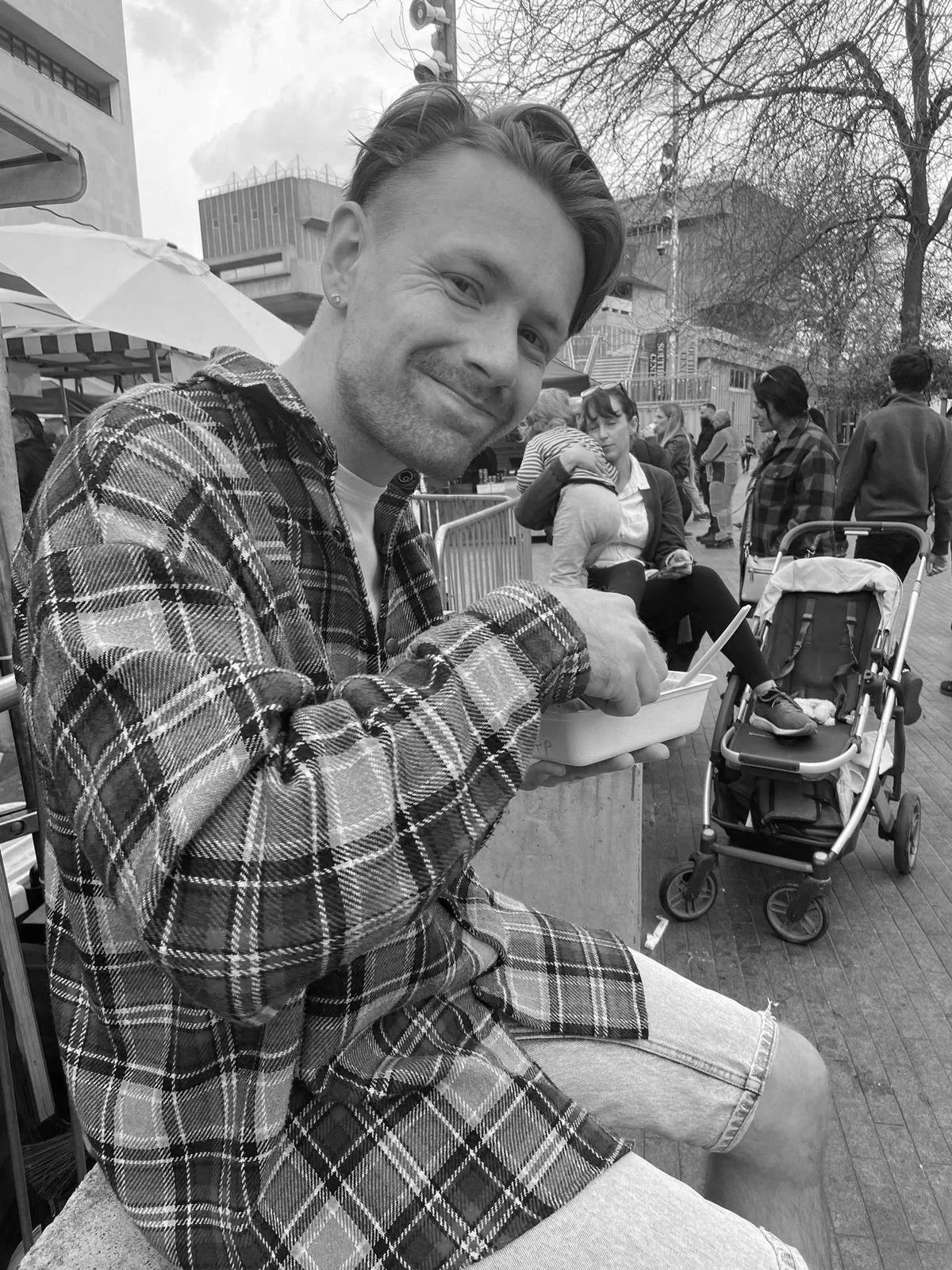 A young man in a plaid shirt sitting outdoors, smiling and holding a container of food, with people and a stroller in the background.