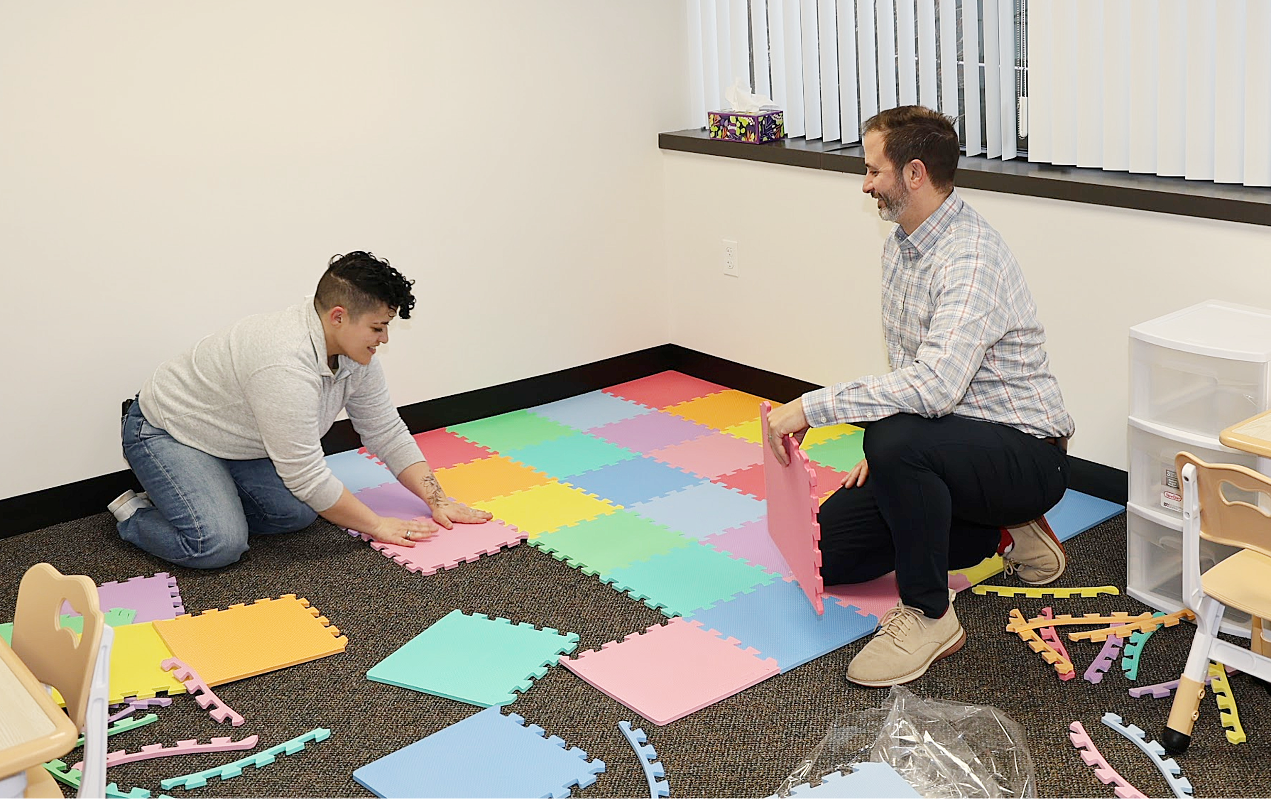 Two people building a colorful foam puzzle floor in a room, one kneeling and one squatting, with foam pieces and a small plastic drawer nearby.
