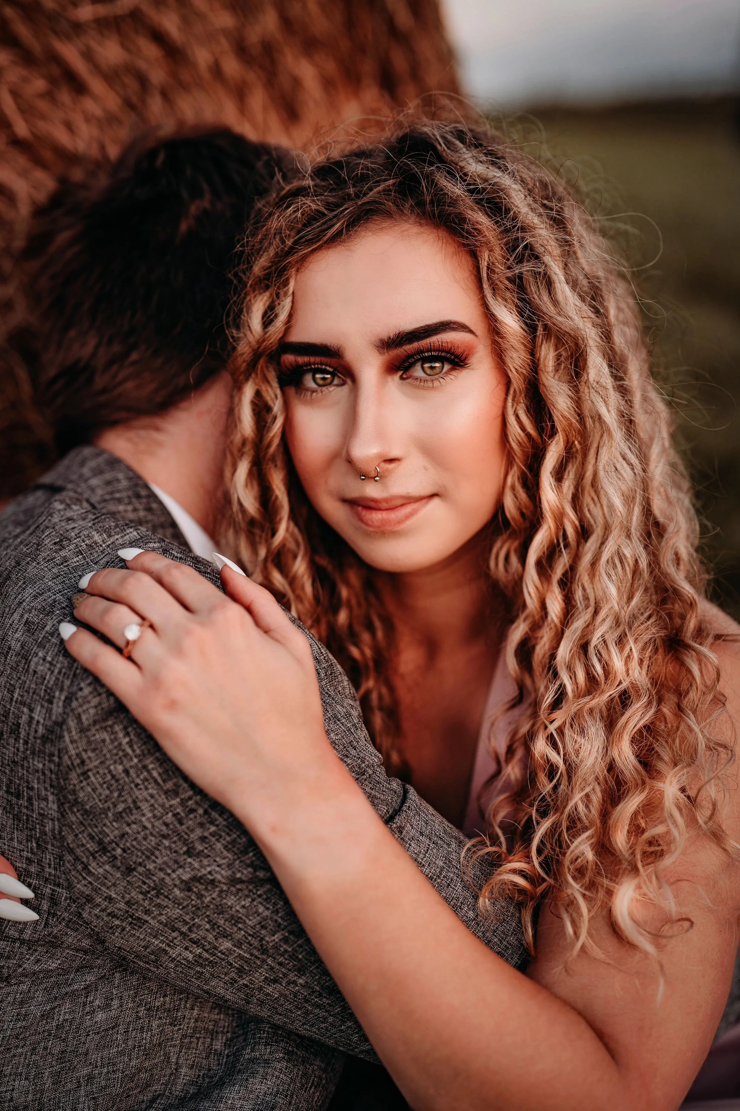 A woman with curly hair and hazel eyes embraces a person wearing a gray suit, with her hand resting on their shoulder, showing a ring and white nails.