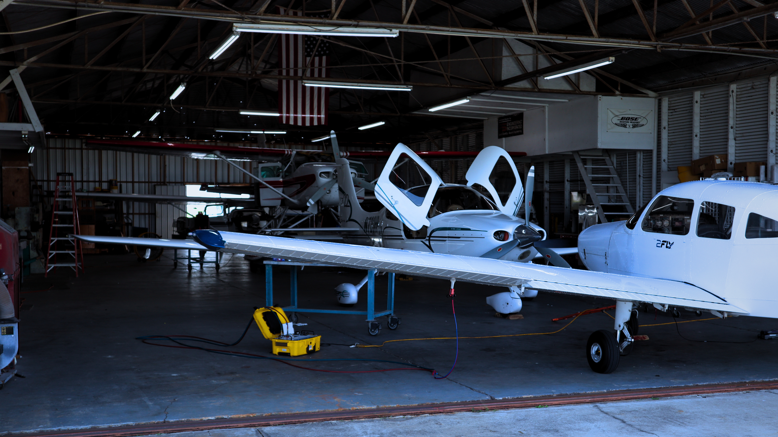 Inside an airplane hangar with two small aircraft, one cirrus with open doors and a partially open fuselage, and a yellow electric toolbox on the ground.