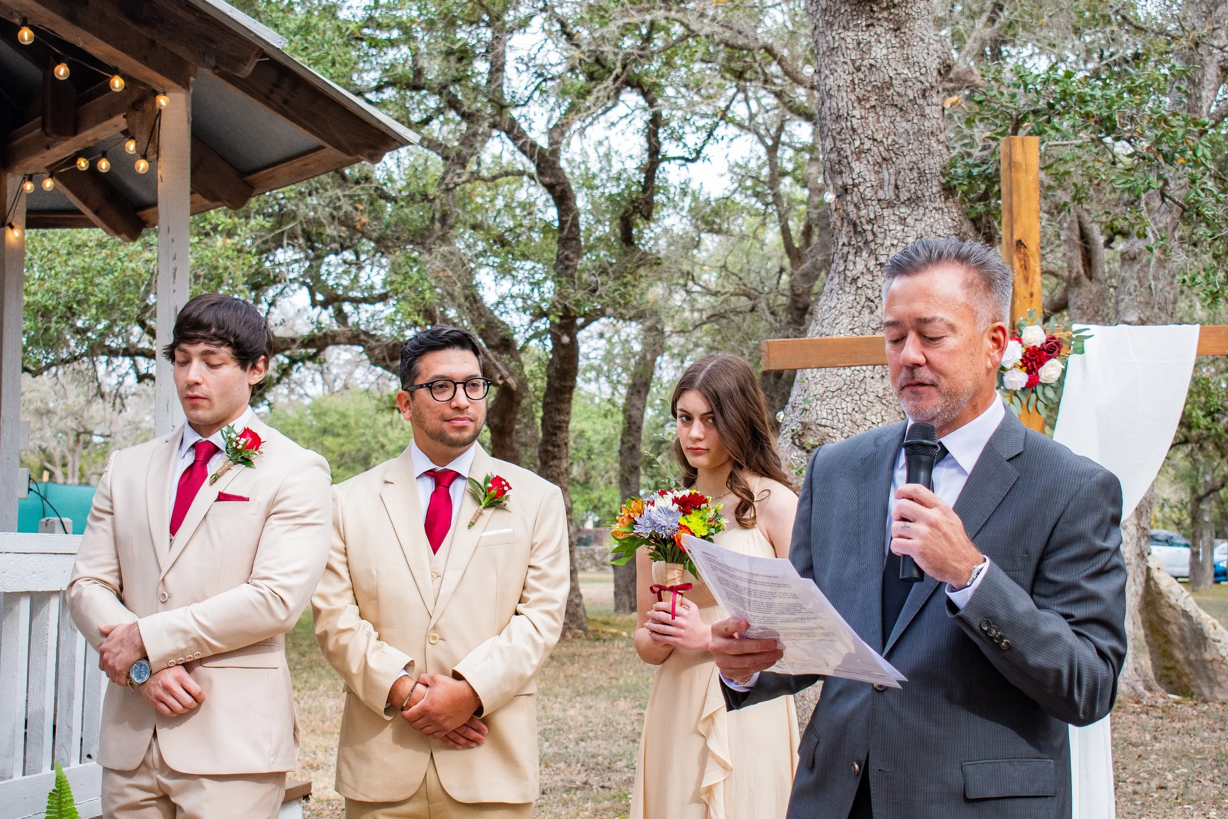 A wedding ceremony outdoors with four people, including two grooms in beige suits with red ties and boutonnieres, a bride in a white dress holding a bouquet, and an officiant in a gray suit reading from a paper. Trees and string lights are in the bac