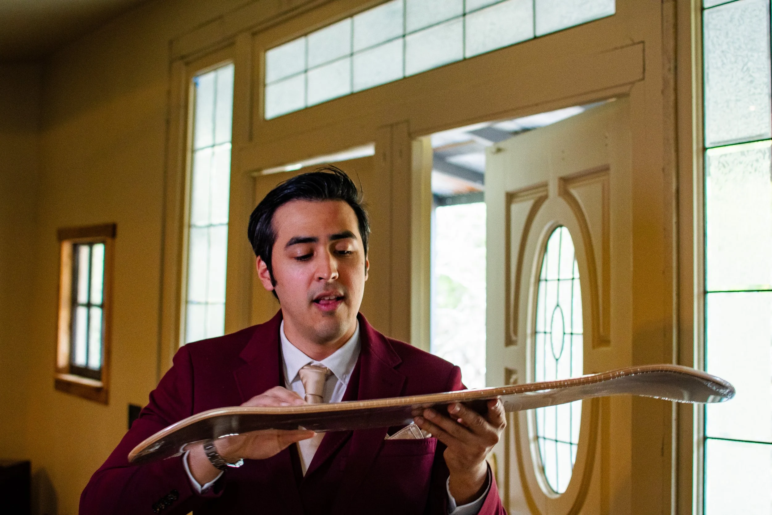 A young man with black hair wearing a burgundy blazer and beige tie looking at a crystal ball inside a house with large windows and a decorative wooden wall.